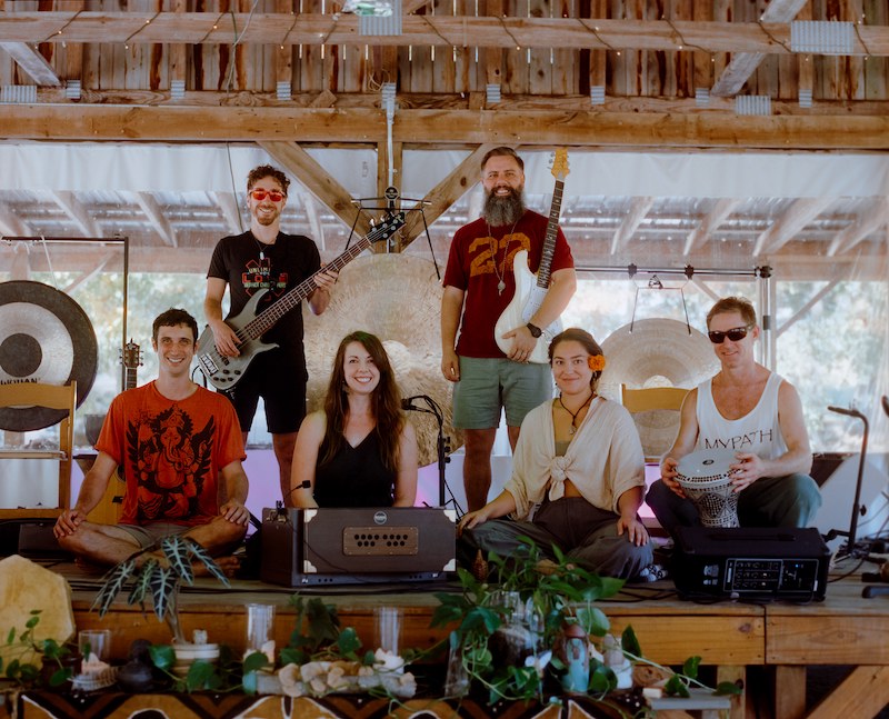 Group of people posing and smiling, holding instruments. Barry stands in the back holding his electric guitar.