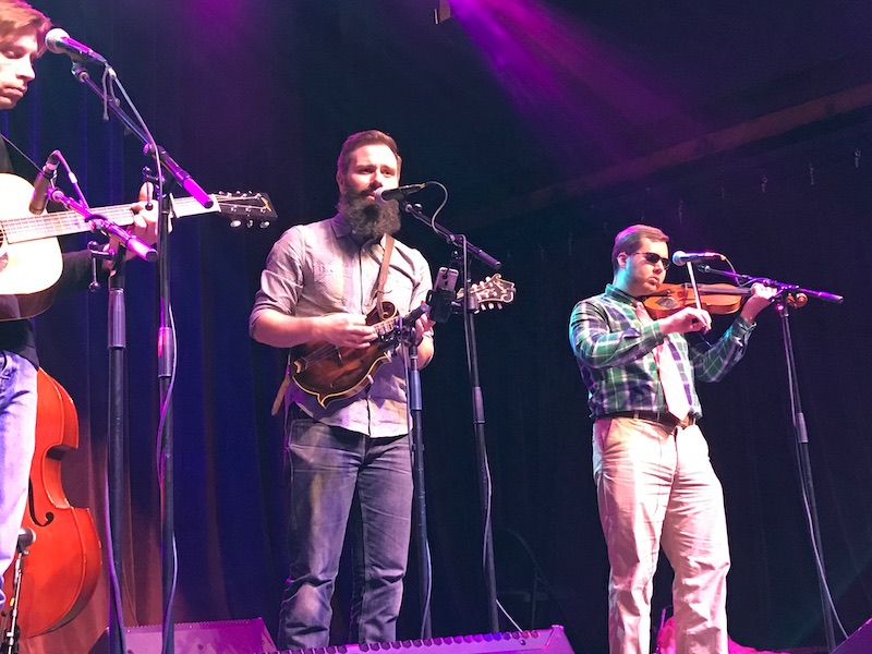 Barry playing mandolin and singing on stage with a fiddle player and guitar player.