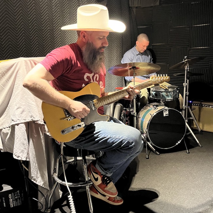 Barry playing a telecaster electric guitar, wearing a large white cowboy hat. Drummer in the background relaxing.